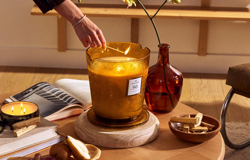 Hand lighting a large Voluspa Baltic Amber jar candle in amber glass, styled with books and dried florals to illustrate luxury candle craftsmanship
